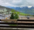 View from a balcony overlooking a parking area, a hotel named "Hotel Sonne," and a scenic mountain landscape with snow-capped peaks and green valleys.