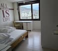 Modern bedroom with a wooden bed, desk, chair, and cabinet, featuring large windows with a scenic mountain view.