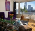 Furnished Room. Modern living room with purple sofa, zebra print rug, glass coffee table, potted plant, and city view through large windows.