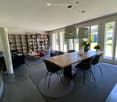 Open-plan living and dining area with large windows, modern furniture, and a bookshelf filled with books.