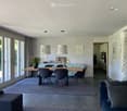 Open-plan living and dining area with a grey sofa, ottoman, dining table with chairs, and a vase with flowers on the table. The space features large windows and a modern aesthetic.