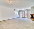 Empty living room with tiled floor, white textured walls, large sliding glass doors leading to outdoor area, fireplace with wooden mantel, and visible kitchen cabinets in the background.