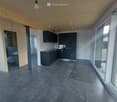 Open-plan space with exposed wooden ceiling beams, grey concrete floor, and large glass sliding doors leading outside. Kitchen area with dark cabinets, sink, and countertop is visible, along with a separate bathroom area with a toilet and sink.