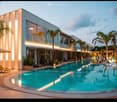 Modern building with a large swimming pool surrounded by lounge chairs, umbrellas, and palm trees.