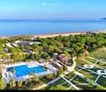 Aerial view of a resort with a swimming pool, lounge chairs, trees, pathways, and a view of the sea and beach.