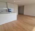 Empty room with light wooden flooring, white walls, and a modern kitchen area with white cabinets and a marble countertop.