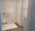Modern bathroom with a white tiled shower area, a sink, and a mirror. The floor is tiled in a neutral color, and the walls are white with a glass shower enclosure.