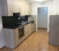 Modern kitchen with light wooden cabinets, stainless steel sink, oven, and a refrigerator. The floor is light wooden, and the walls are white with a black backsplash.