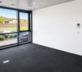 Empty room with white walls, grey carpet, large glass sliding doors, and a view of a parking lot and greenery.