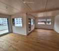 Empty room with wooden flooring, white walls, and large windows offering views of the city and surrounding buildings.