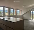 Modern, unfurnished kitchen with sleek, grey cabinetry, a central island, and large windows offering a scenic mountain view. The space features a wooden floor and recessed lighting.