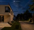 Modern two-story house with a raised ground floor, illuminated entrance steps, and a balcony on the upper level.