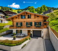A two-story wooden house with green shutters, a balcony, and a garage, situated in a lush, green landscape with a raised ground floor and a view of the surrounding hills.