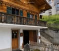 Traditional wooden house with green shutters, a balcony, and a small garden area. The house features a steeply pitched roof and a stone staircase leading up to the entrance.