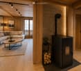 Modern living room with wooden walls and ceiling, a white sectional sofa, a round coffee table, a wood-burning stove, and large windows with curtains.