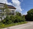 Two-story house with white exterior, large windows, balcony, and solar panels on the roof, surrounded by greenery.