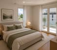 Modern bedroom with a large bed, nightstands, lamps, a picture frame, and a view of the landscape through large windows and sliding glass doors leading to a balcony.