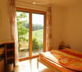 Bright bedroom with wooden floor, large sliding glass doors leading to a garden, a bed with a colorful duvet, a nightstand, and a cabinet with shelves.