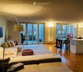 Open-plan living and dining area with a sectional sofa, a round wooden stool, a dining table with chairs, a modern kitchen island, large sliding glass doors leading to a balcony, and a floor lamp.