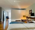 Modern bedroom with two single beds, a desk with a chair, a lamp, and a screen. The room features wooden flooring and large windows.