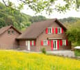 Two-story brown house with red shutters and a tiled roof, surrounded by a lush green landscape with yellow flowers in the foreground.