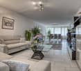 Modern living room with a beige sofa, glass coffee table, dining area with chairs and table, television, and decorative plants.