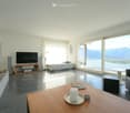 Modern living room with large windows offering a scenic view, featuring a sofa, coffee table, ottoman, armchair, TV, floor lamp, and fireplace.