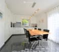 Modern kitchen with white cabinets, a wooden dining table, black chairs, and a bowl of fruit on the table. The space features a sleek design with a large window and a hanging light fixture.