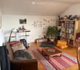 Living room with a couch, coffee table, rug, bookshelves, potted plant, desk, stools, and various decorative items.
