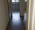A narrow hallway with dark flooring, leading to a small kitchen area visible at the end. The kitchen has white cabinets, a sink, and a window above it.