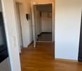 A well-lit hallway with wooden flooring, white walls, and a partially open door leading to another room. The hallway features a light switch and a small intercom system mounted on the wall.
