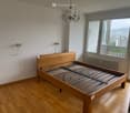 Spacious bedroom with wooden floor, large window offering scenic view, wooden bed with metal slats, chandelier, radiator, and electrical outlets.