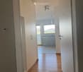 A narrow hallway with wooden flooring, white walls, and built-in cabinets on one side. A view into a kitchen area with white cabinets and a dishwasher is visible at the end of the hallway.