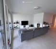 Modern living room with grey sectional sofa, coffee table, TV, shelves, and a view through large windows.