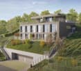 Modern two-story house with a flat roof, large windows, and a balcony on the upper level. The house is situated on a hillside with a landscaped garden and a covered garage at the base.