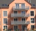 A modern, three-story apartment building with orange facade and multiple balconies. The ground floor features a small garden area with outdoor furniture.