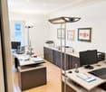 Modern office space with two desks, computer monitors, keyboards, mice, a floor lamp, cabinets, and framed artwork on the walls.
