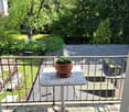 Balcony with a small table and potted plant, overlooking a green garden area with trees and a building in the background.