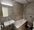 Modern bathroom with beige tiles, a white sink, bathtub, toilet, and a large mirror with built-in lighting. Features a handheld showerhead and grab bars for safety.