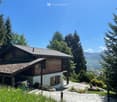 A two-story wooden house with a tiled roof, surrounded by trees and mountains, with a driveway and garden area.