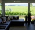 Living room with large windows overlooking a green field and mountains, featuring a dark leather sectional sofa with pillows and stuffed toys, a small table, potted plants, and a picture on the wall.