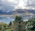 A scenic outdoor view featuring a lush garden with various trees and shrubs in the foreground, overlooking a serene lake surrounded by mountains and a town under a partly cloudy sky.