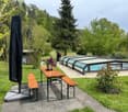 Garden area with a wooden table, benches, a covered patio, and a swimming pool surrounded by greenery.