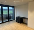 Modern, minimalist room with light wooden flooring, large glass sliding doors leading to a balcony with scenic countryside view, a sleek black desk with drawers, and a tall white wardrobe.