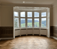 Empty room with large bay windows offering a scenic view of a green landscape. The room features wooden flooring and decorative wall paneling.
