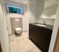 Modern bathroom with a toilet, sink, and large mirror. Features a window with a view of greenery, tiled floor, and sleek dark cabinetry.