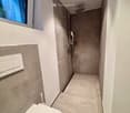 Modern bathroom with a toilet, shower, and a window providing natural light. The walls are tiled in neutral tones, and the floor is covered with light-colored tiles.