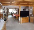 A spacious, well-furnished living and dining area with wooden beams on the ceiling, a large flat-screen TV, a dining table with chairs, and wooden cabinets. Shelves and decorative items are visible along the walls.