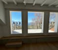 Empty room with wooden flooring, white walls, and three large windows offering a scenic view of the ocean and mountains. A small wooden bench is positioned near the windows.