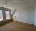 Empty room with wooden flooring, white walls, large windows, built-in cabinets, radiator, and exposed wooden beams on the ceiling.